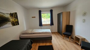 Ein Schlafzimmer mit Bett, einem Fenster mit Vorhängen, einem Sessel, einem TV-Schrank und einem Bild an der Wand.