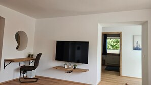 A modern living room with a flat-screen TV mounted on the wall, a wooden desk with a chair, and a small shelf with decorative items.