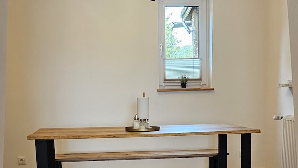A room with a wooden bench, a table, a hanging light fixture, and a window with blinds.