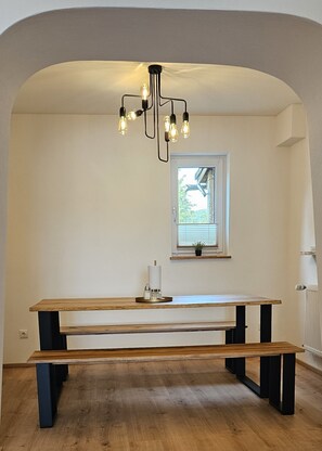 Een kamer met een houten bank, een tafel, een hanglamp en een raam met jaloezieën.