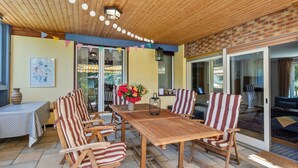 A covered patio with a wooden table and striped chairs, a vase with flowers, and a view through glass doors.
