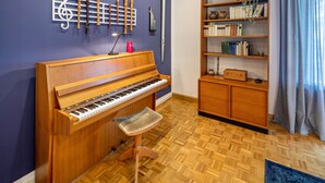 A room with a wooden piano, a bookshelf, and a wall-mounted music stand with instruments.