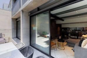 A modern living room with a glass door leading to a balcony, a kitchen area, and a seating area with a sofa and chairs.