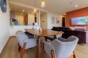 Een moderne woonkamer met een eetgedeelte, een paarse bank en een televisie waarop een kamer te zien is.