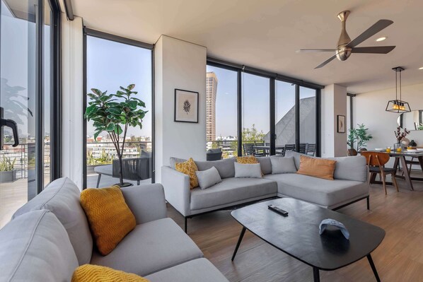 Un salon moderne avec un canapé d’angle gris, une table basse noire et un ventilateur de plafond. On y trouve également un coin repas avec une table et des chaises, ainsi qu’une grande fenêtre offrant une vue sur la ville.