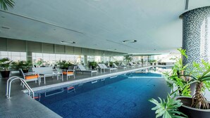 A modern indoor pool with clear glass walls, lounge chairs, and potted plants.