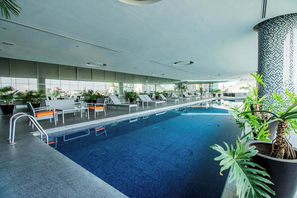 A modern indoor pool with clear glass walls, lounge chairs, and potted plants.