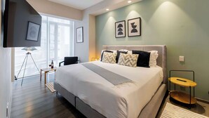 A modern hotel room with a large bed, a flat-screen TV mounted on the wall, a floor lamp, and a small table.