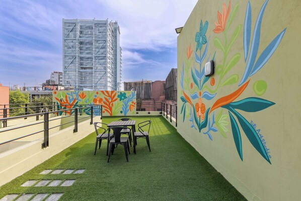 Un patio exterior con césped artificial, una mesa y sillas, y un mural colorido en la pared.