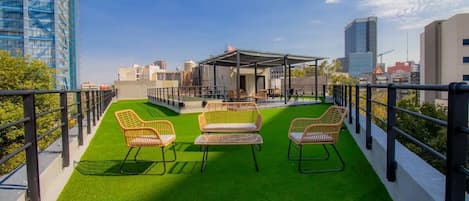 A rooftop terrace with green artificial grass, wicker chairs, and a covered seating area.