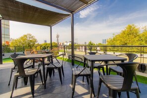 Une terrasse extérieure avec des tables et des chaises, un espace abrité et une vue sur les bâtiments et les arbres.
