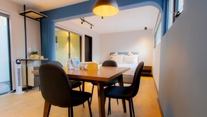 Ein modernes Hotelzimmer mit einem Holztisch, vier Stühlen, einem Bett mit weißer Bettwäsche, einem Nachttisch mit Lampe und einem großen Fenster.