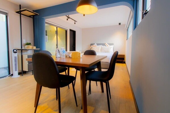 A modern hotel room with a wooden dining table, four chairs, a bed with white bedding, a bedside table with a lamp, and a large window.