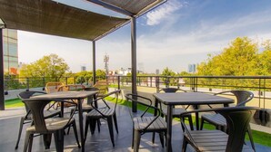 Eine Terrassensitzgruppe mit Tischen und Stühlen, ein überdachter Bereich und Blick auf Gebäude und Bäume.