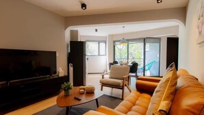 Ein modernes Wohnzimmer mit einer Couch, einem Couchtisch und einem Fernseher. Ein Sessel und ein kleiner Tisch mit einer Pflanze stehen ebenfalls im Raum. Große Fenster bieten einen Blick ins Freie.