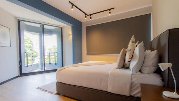 Ein Schlafzimmer mit einem großen Bett, einem dunklen Kopfteil, einem Nachttisch mit Lampe und Blick durch eine Glastür auf einen Balkon.