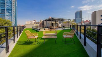 A rooftop terrace with green artificial grass, wicker chairs, and a covered seating area.