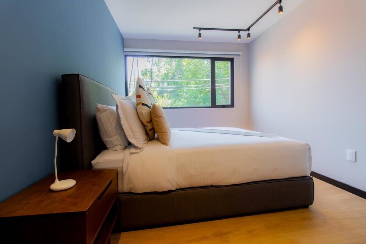 A modern bedroom with a bed, a bedside table, a lamp, and a window with a view of greenery.