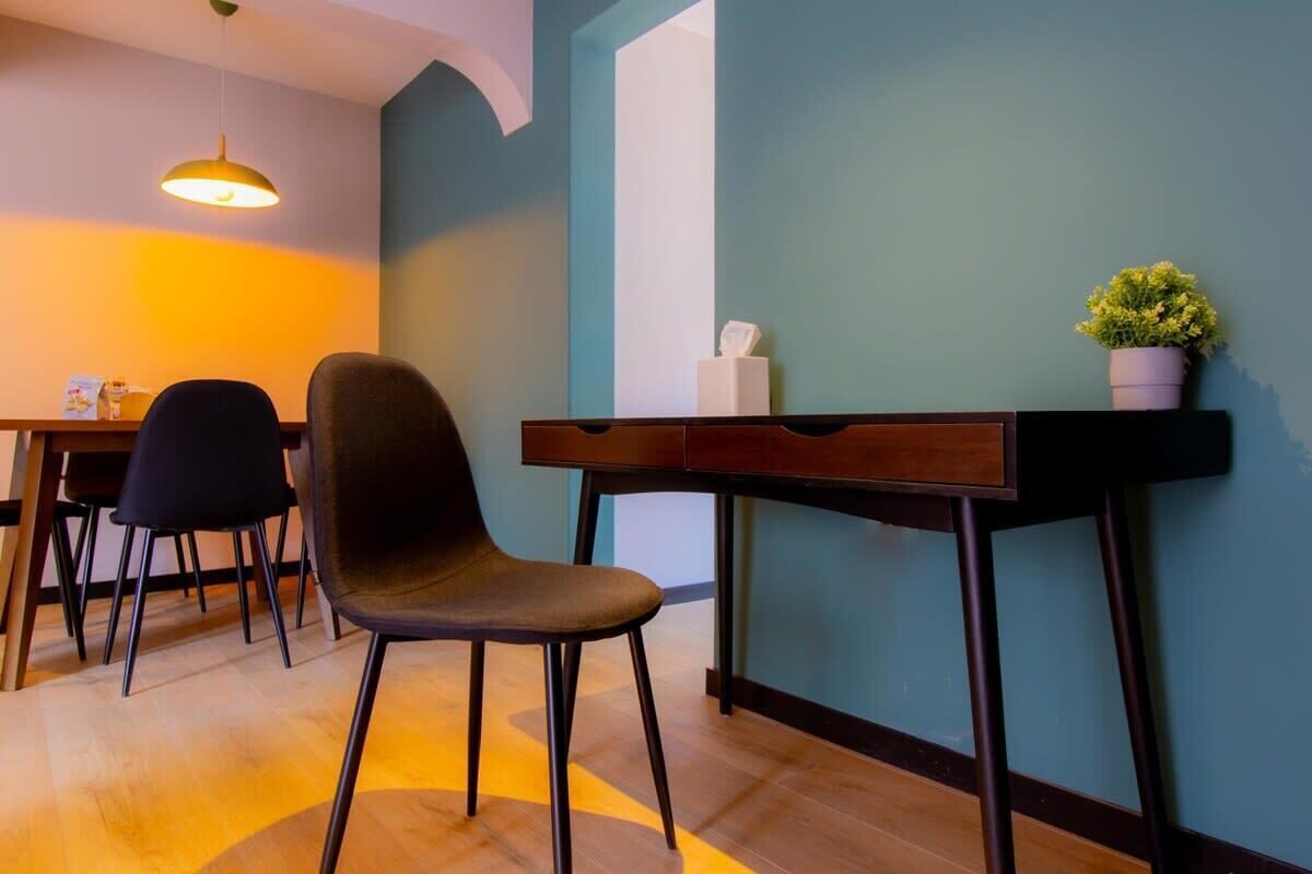A modern room with a wooden floor, a desk with a chair, a hanging light, and a plant on the wall.