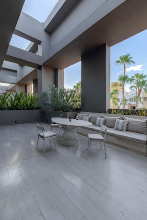 An outdoor patio area with a round table, chairs, a sofa, and a large window overlooking a cityscape.