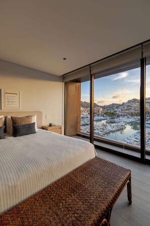 A hotel room with a large bed, a wooden bench, a nightstand, and a view of a marina and cityscape.