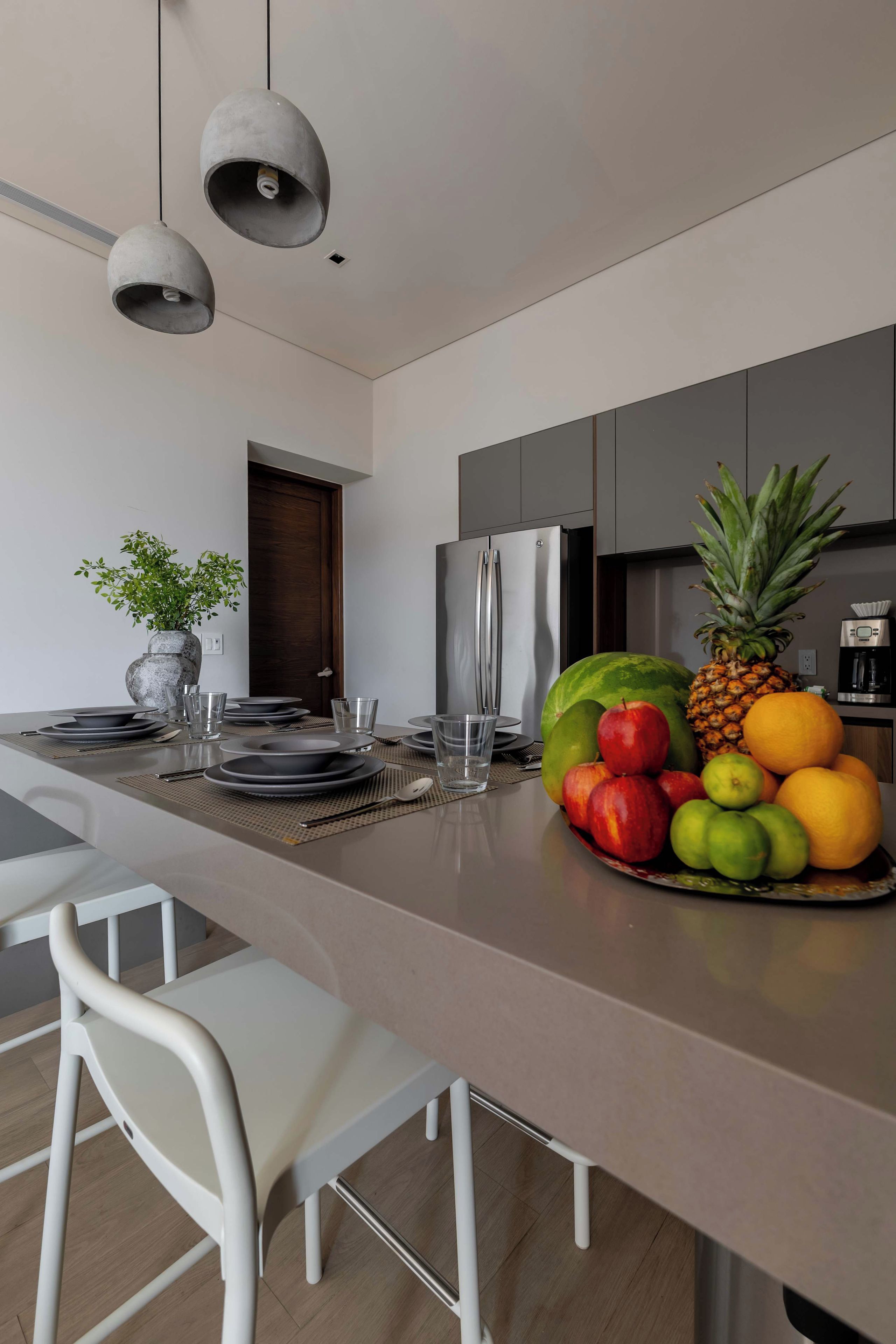 A modern kitchen with a central island, white chairs, and a fruit bowl.