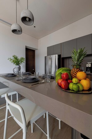 A modern kitchen with a central island, white chairs, and a fruit bowl.