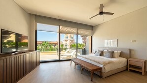 Une chambre avec un lit, un banc, une télévision, un ventilateur de plafond et une vue sur un bâtiment et de la verdure à l’extérieur.