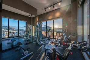 Ein Fitnessstudio mit unterschiedlichen Trainingsgeräten, großen Fenstern und Blick auf eine Stadtlandschaft.