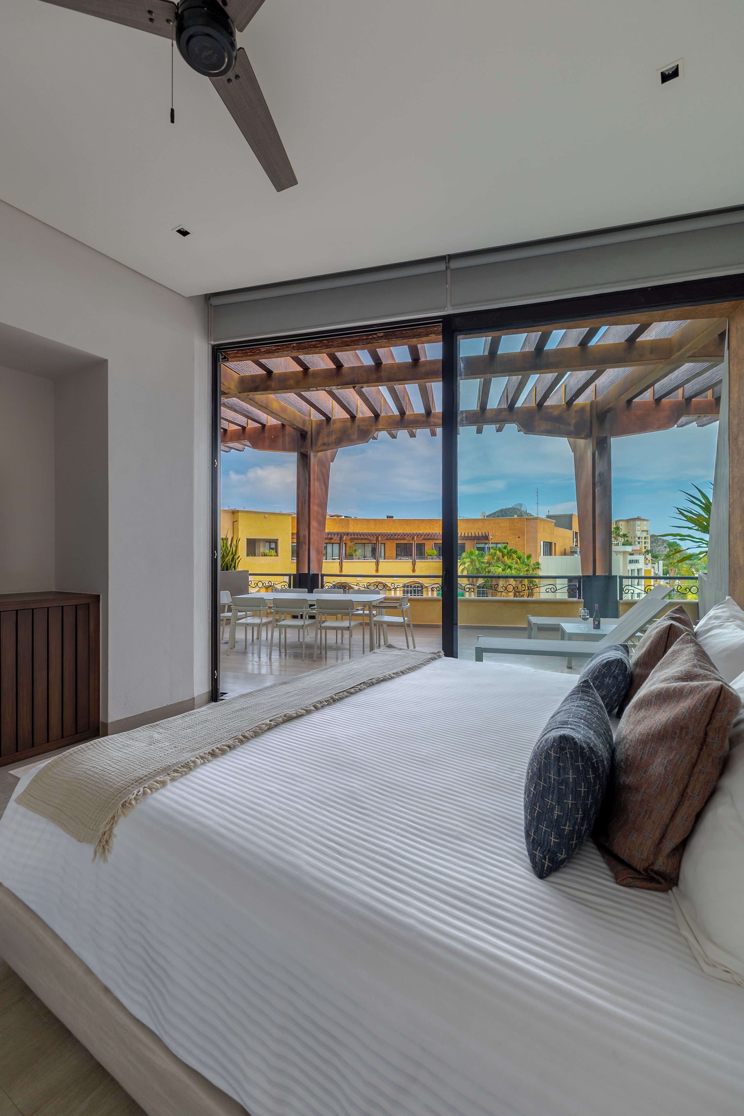 Quarto moderno com cama grande, ventilador de teto e vista para uma varanda com mobiliário de exterior.