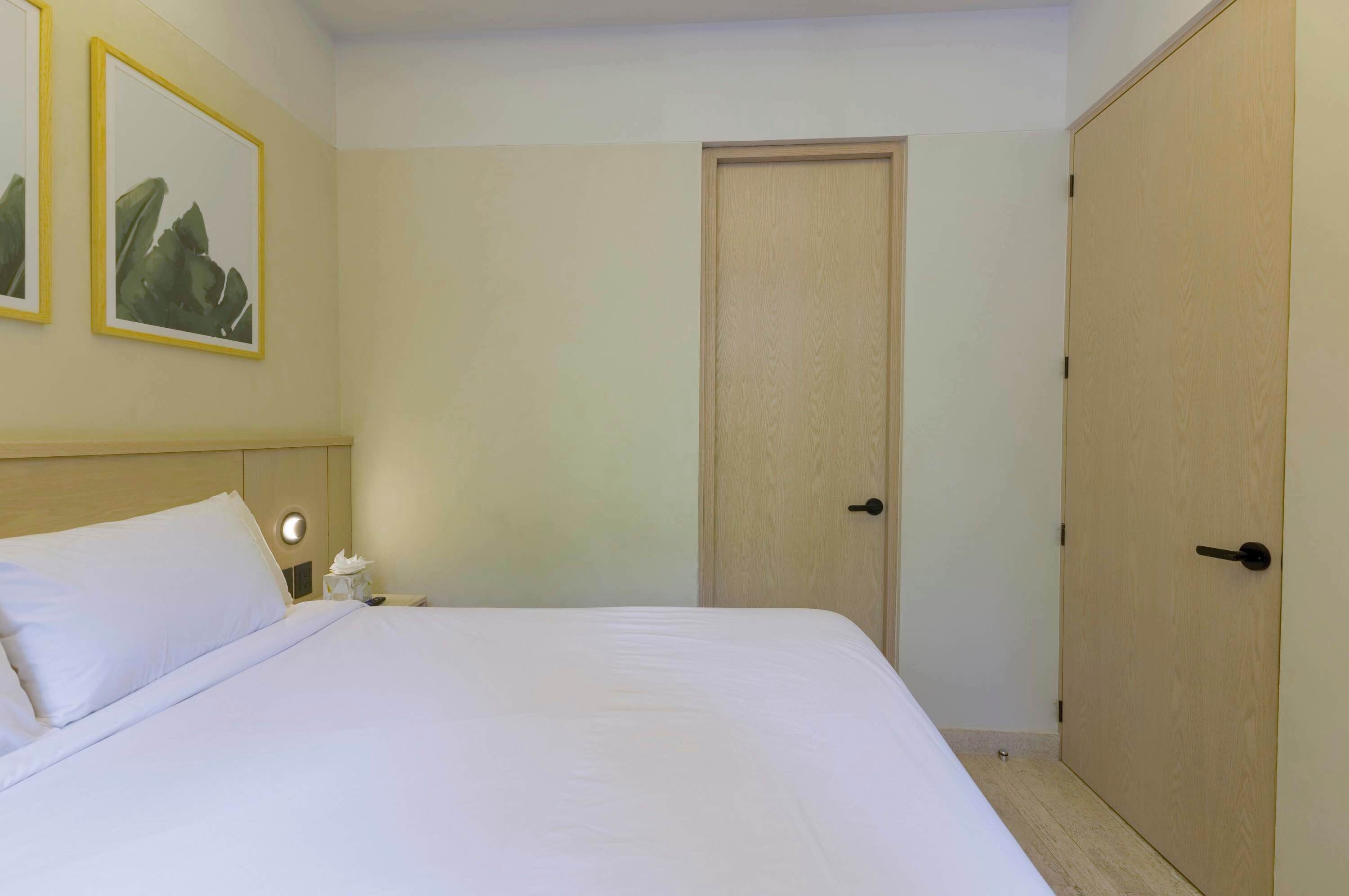 A hotel room with a bed, a bedside table, a nightstand, a door, and two framed pictures on the wall.