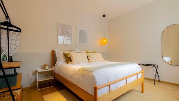 A bedroom with a wooden bed, a nightstand, a hanging light, and a mirror on the wall. - Capitalia | Chill Stay & Near Condesa & Museums (Ciudad de México)