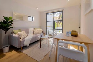 Uma sala de estar moderna com sofá, mesa de jantar e cadeiras. Há uma janela grande que oferece luz natural e vista para árvores.