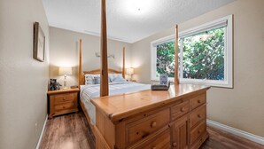 Een slaapkamer met een grote houten kapstok, een hemelbed, een nachtkastje met een lamp en een raam met uitzicht op bomen.