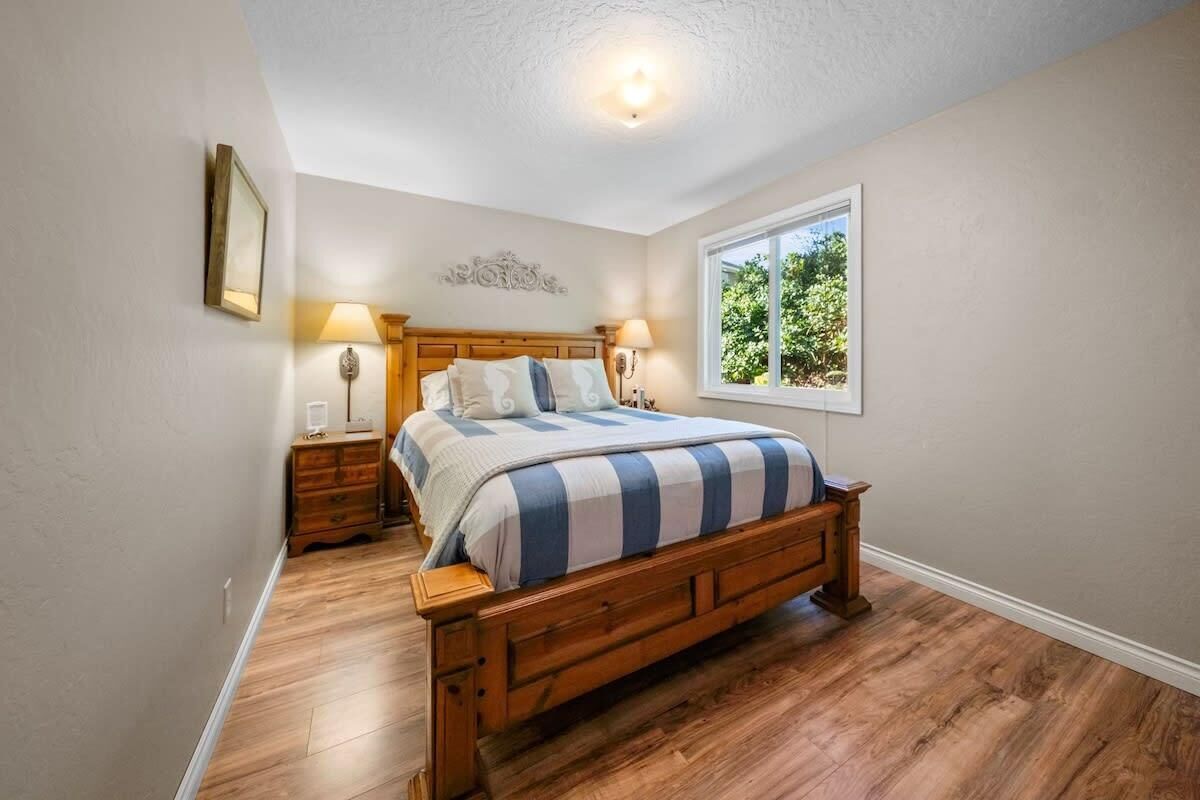 A bedroom with a wooden bed, two bedside tables, a nightstand, a window with a view of greenery, and a ceiling light.