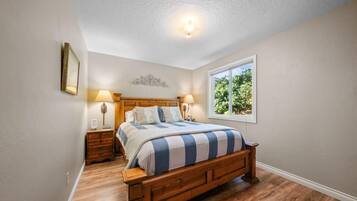 A bedroom with a wooden bed, two bedside tables, a nightstand, a window with a view of greenery, and a ceiling light.