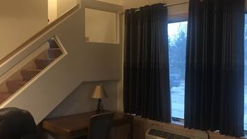 A living room with a staircase, a leather sofa, a table with a lamp, and a window with curtains.