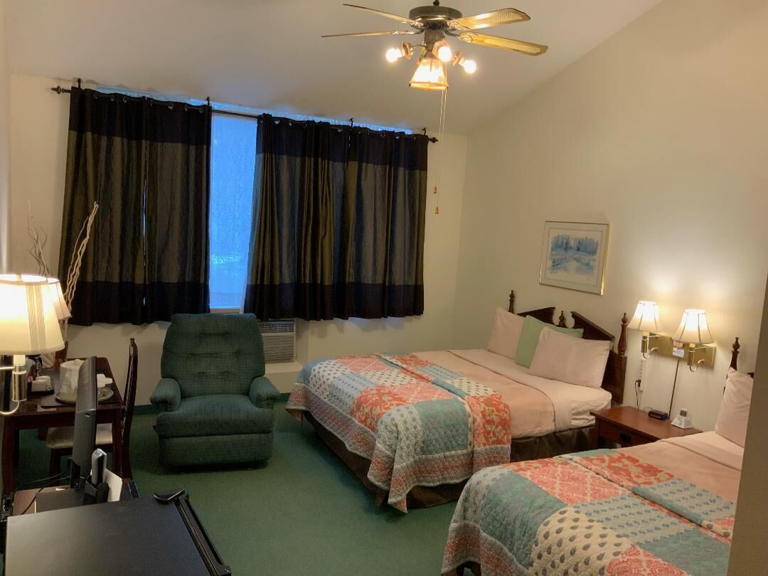 Habitación de hotel con dos camas, un escritorio, una silla, un ventilador de techo y una ventana con cortinas.