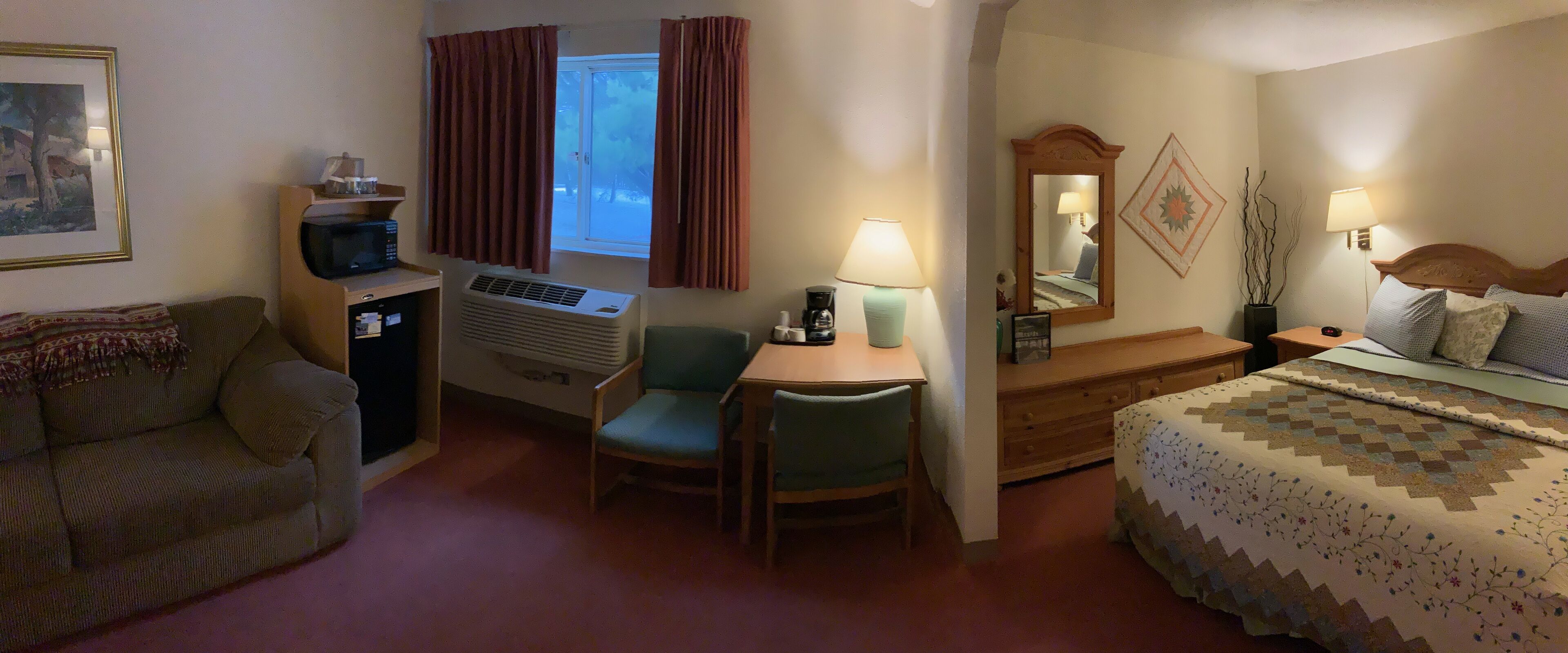 Quarto de hotel com cama, escrivaninha com cadeira, micro-ondas, aquecedor, janela com cortinas, espelho e luminária.