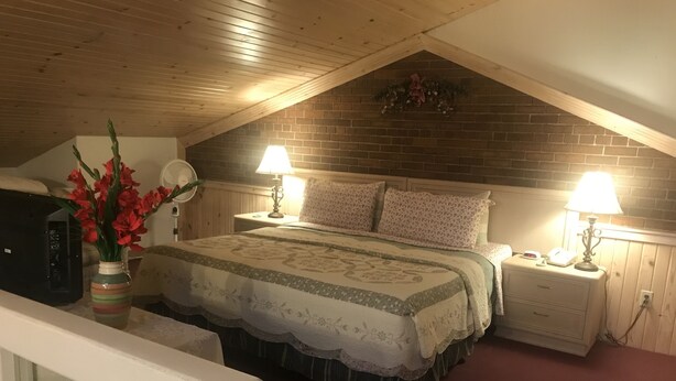 A bedroom with a sloped ceiling, a brick wall, a bed with floral bedding, two bedside lamps, and a vase with red flowers. - Country Lodge inn (Harmony)