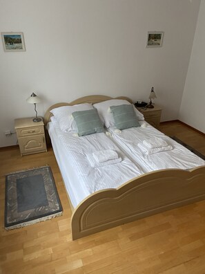 Una cama bien tendida con sábanas blancas y dos almohadas, una mesita de noche con una lámpara, una pequeña alfombra y dos cuadros en la pared.
