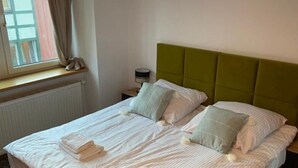A neatly made bed with white linens and two folded towels, a green upholstered headboard, and a window with curtains. - Kauris Apartament (Gdańsk)