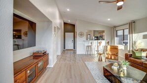 A spacious living area with a wooden cabinet, a coffee table, and a sofa. There is a kitchen area in the background with a bar and stools. - Mid Century Themed Home on the Golf Course (San Tan Valley)