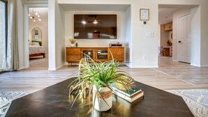 A modern living room with a dark coffee table, a potted plant, and a book. A wooden cabinet and a large flat-screen TV are visible in the background. - Mid Century Themed Home on the Golf Course (San Tan Valley)