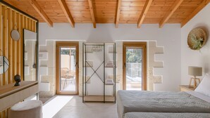 A bedroom with a wooden ceiling, a bed, a nightstand, a mirror, and a view through a glass door to an outdoor seating area. - Fillio luxury villa (Αδελιανός Κάμπος)