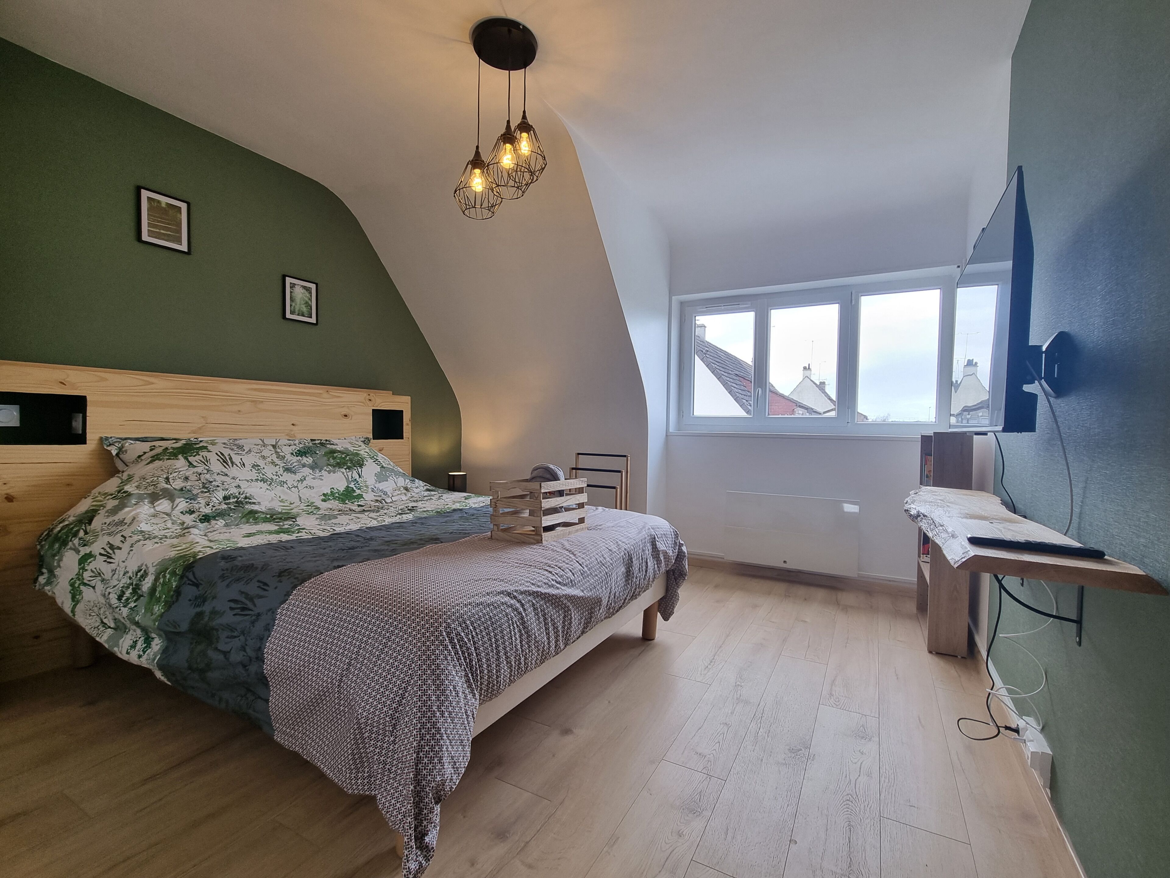 A bedroom with a sloped ceiling, a bed with floral bedding, a wooden headboard, a bedside table, and a window with a view of buildings.