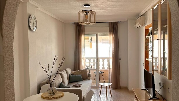 A modern living room with a sofa, dining table, and chairs, a TV, and a balcony with curtains. - Casa de Luz - Beach & Boulevard (Torrevieja)