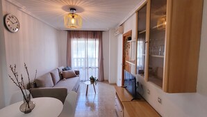 A modern living room with a sofa, a dining table with a vase, a TV, and a large window with curtains. - Casa de Luz - Beach & Boulevard (Torrevieja)