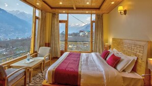 A hotel room with a large bed, a wooden ceiling, a view of mountains, and a balcony with a table and chair. - Sparklyn View (Manali)