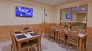 A dining area with wooden furniture, a television mounted on the wall, and a large window. - Sparklyn View (Manali)